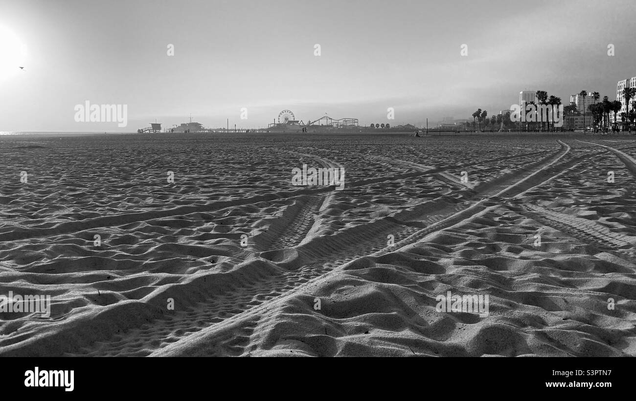 Vista in bianco e nero sulla spiaggia di Santa Monica con molo, hotel e appartamenti in lontananza. Concentrarsi sui cingoli degli pneumatici nella sabbia Foto Stock