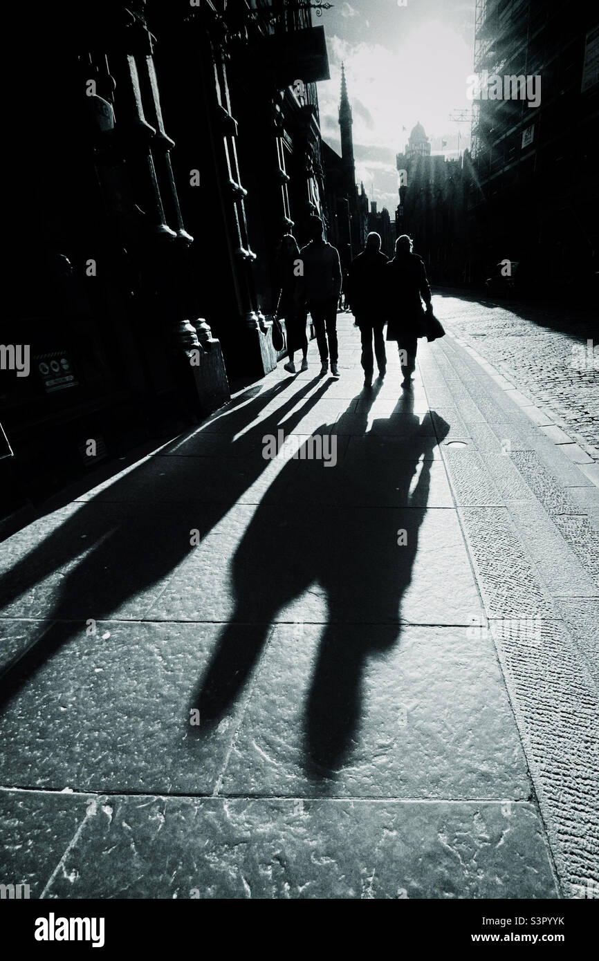 Le persone che camminano lungo il Royal Mile a Edimburgo sono retroilluminate dal sole nel tardo pomeriggio e gettano lunghe ombre Foto Stock