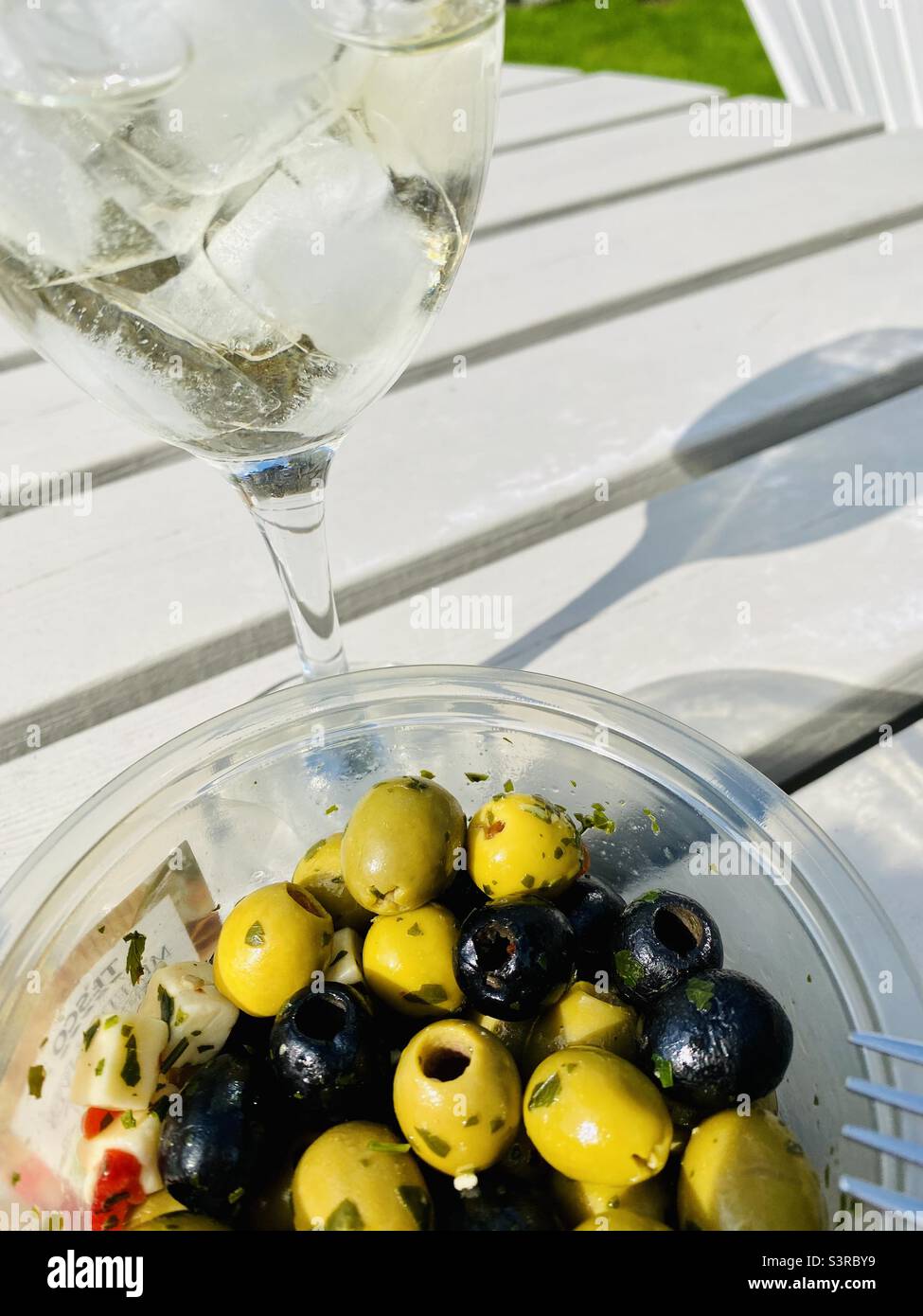Vino bianco con ghiaccio e una ciotola di olive verdi e nere sul tavolo da giardino al sole - uno spuntino estivo perfetto Foto Stock
