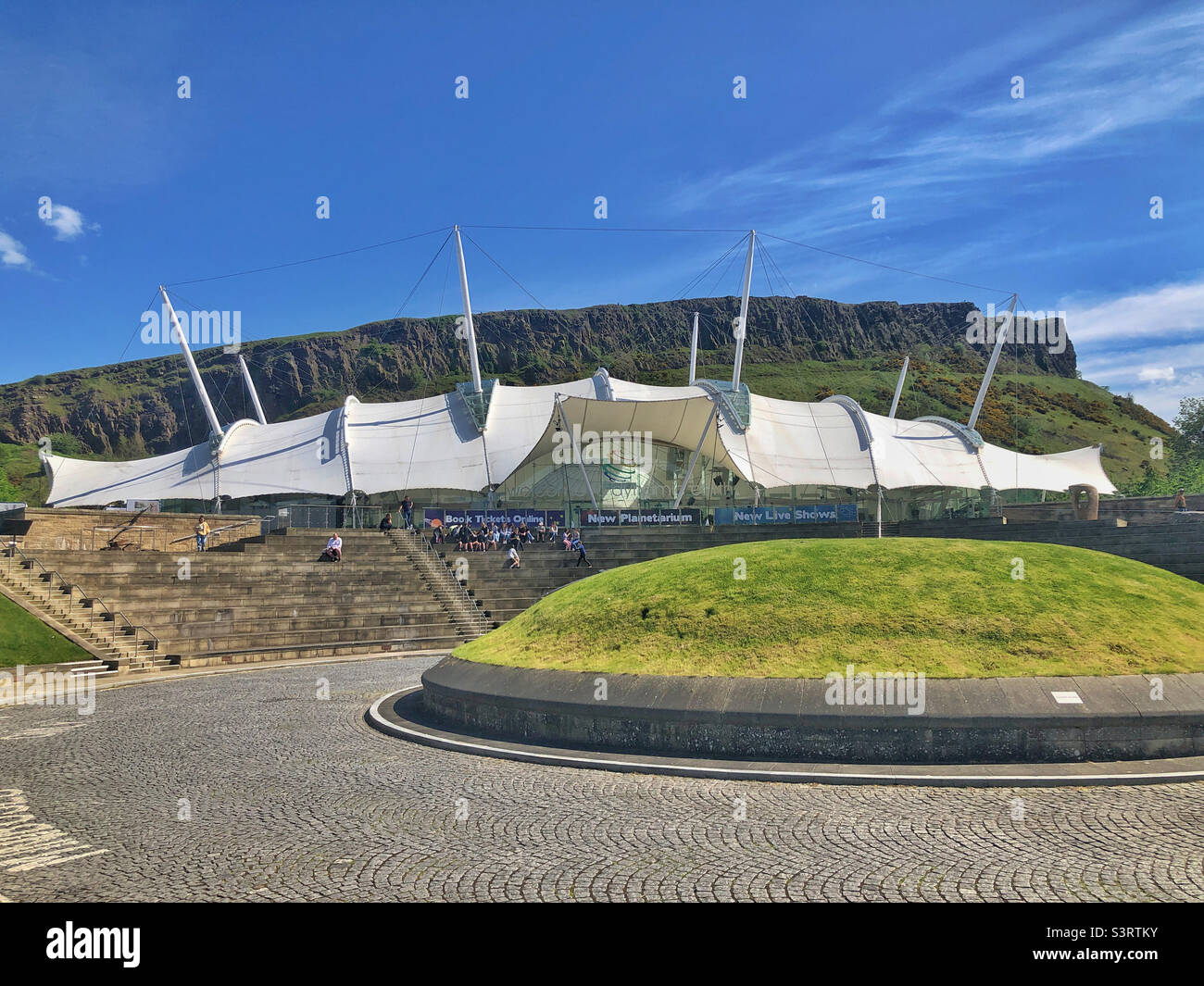 Dynamic Earth, Edimburgo, Scozia, in una giornata di sole. Attrazione per famiglie all'avanguardia che porta i visitatori attraverso la storia dal Big Bang ai giorni nostri. Salisbury Crags è sullo sfondo. Foto Stock