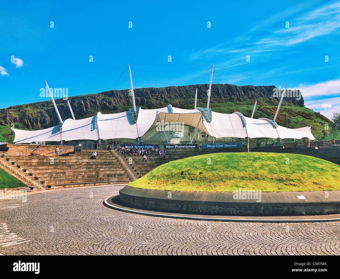 Dynamic Earth, Edimburgo, Scozia in una giornata di sole. Attrazione per famiglie all'avanguardia che porta i visitatori attraverso la storia dal Big Bang ai giorni nostri. Salisbury Crags è sullo sfondo. Foto Stock