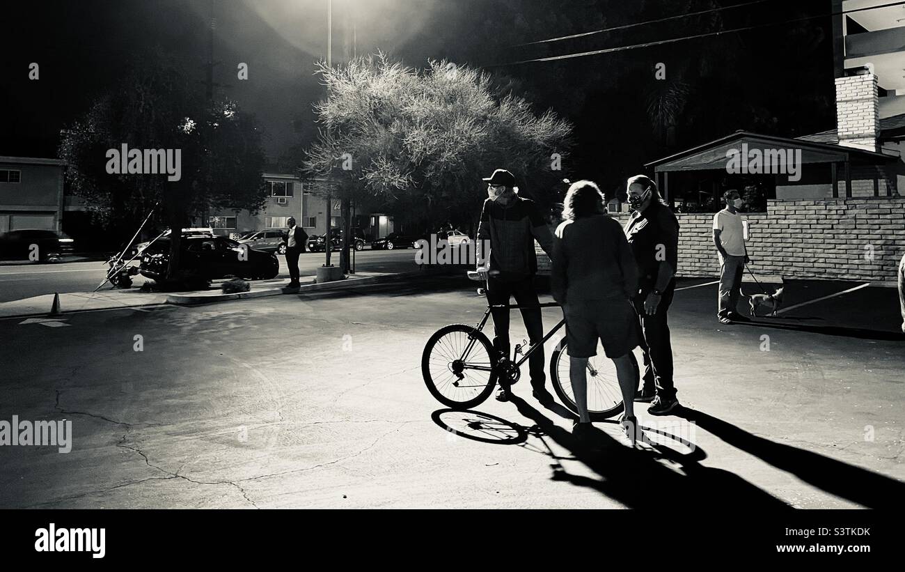 LOS ANGELES, CA, Oct 2021: Tre uomini, uno con una bicicletta, con una vista dalla luce della strada mentre si trova in un parcheggio di notte. Bianco e nero Foto Stock