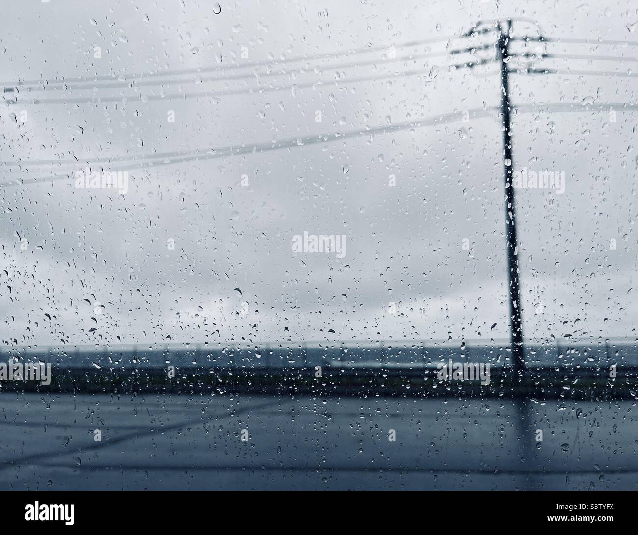 Gocce di pioggia sulla finestra di vetro che guarda verso l'asta di elettricità in un parcheggio vicino al mare - Immagine stock catturata con smartphone