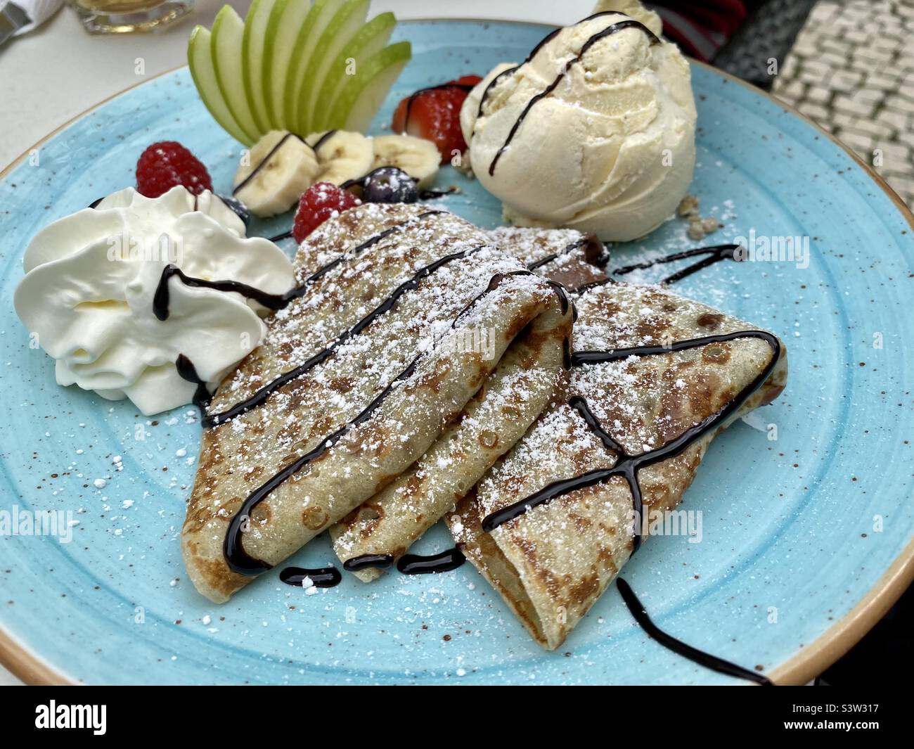 Piatto blu con crepes al cioccolato, frutta fresca e un cucchiaio di gelato alla vaniglia Foto Stock