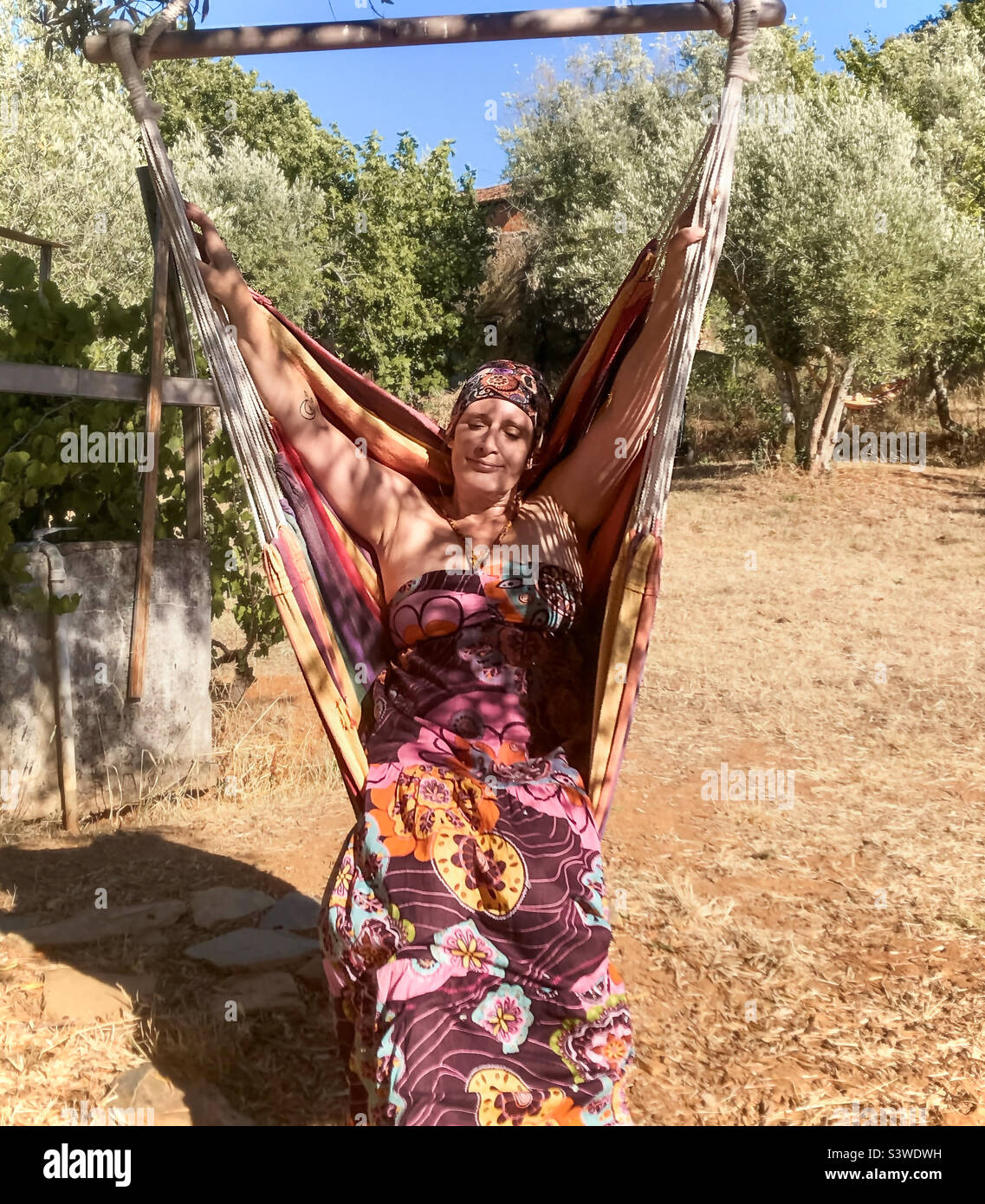 Donna si rilassa sotto l'ombra di un albero in una sedia di amaca swing, mentre indossa un abito colorato maxi - Immagine stock catturata con smartphone