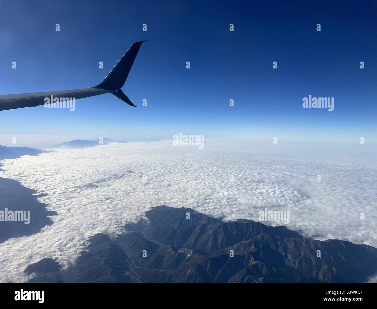 Vista dall'aereo di linea commerciale con ala dell'aeromobile, vista dalla finestra dell'aereo mentre sorvola le montagne che si innalzano sopra le nuvole basse Foto Stock