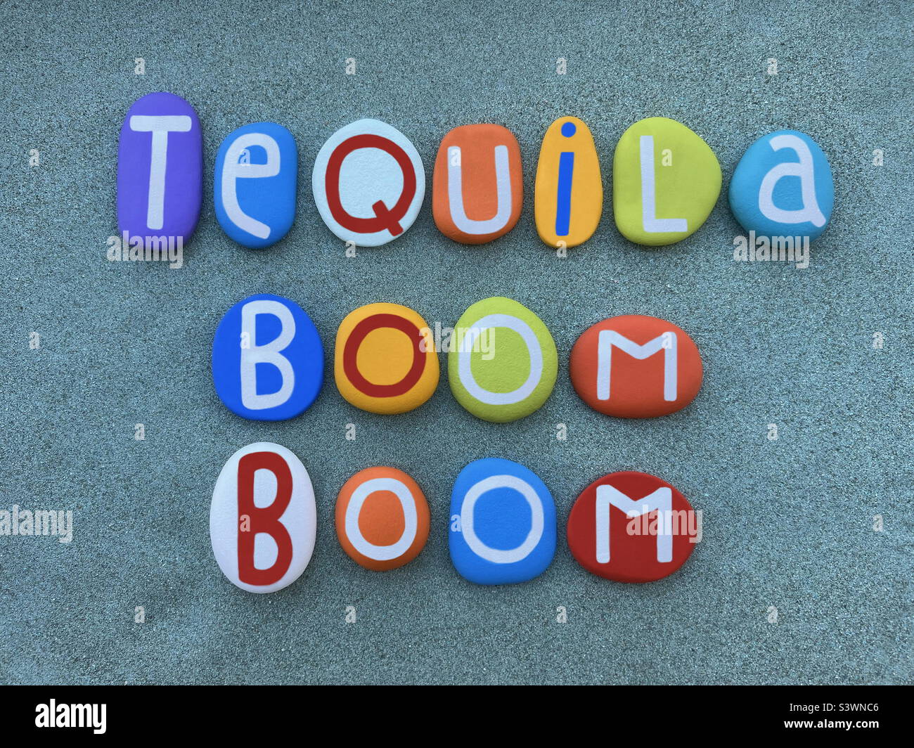 Tequila boom, testo creativo del nome del cocktail composto da lettere in pietra multicolore dipinte a mano su sabbia verde - Immagine stock catturata con smartphone