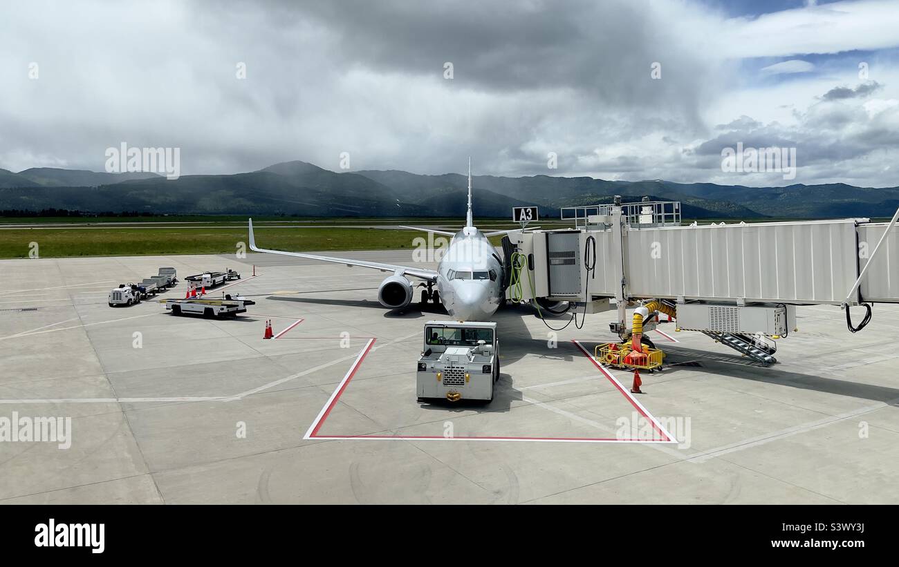 MISSOULA MONTANA AIRPORT, MT, GIUGNO 2022: Vista ampia American Airlines Embraer E175 jet velivolo parcheggiato al cancello del terminal con veicoli a terra intorno ad esso, montagne sullo sfondo, nessun logo visibile - Immagine stock catturata con smartphone