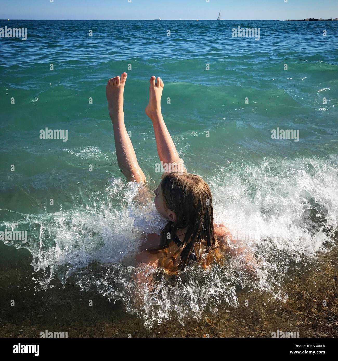 Una ragazza seduta sul bordo dell’acqua sulla spiaggia di Barcellona in una vacanza in famiglia che permette alle onde di schiantarsi su di lei e sollevarsi le gambe di gioia Foto Stock