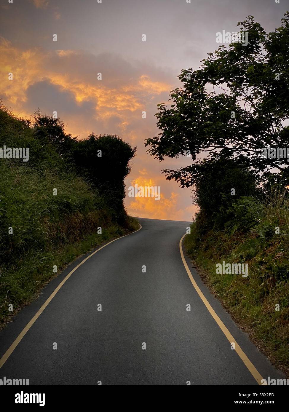 Una tradizionale strada di campagna britannica con siepi che conducono su una collina ad un cielo scintillante e paradisiaco al tramonto Foto Stock
