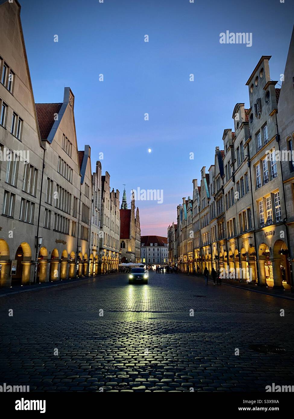 Una sola auto guida giù per una strada acciottolata al crepuscolo nella città vecchia della città tedesca di Munster. Oscurità ai margini della città. Nel retrogusto della luna del tramonto. Come una scena da un film spia. Foto Stock