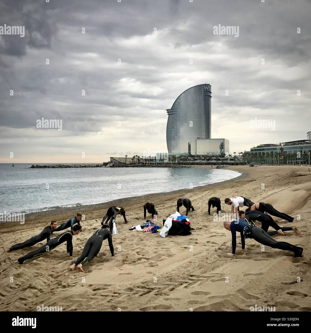 Una scuola di surf studenti si scaldano per la loro lezione mattutina sulla trafficata spiaggia della città di Barceloneta, di fronte al W Barcelona Hotel a Barcellona, Spagna Foto Stock