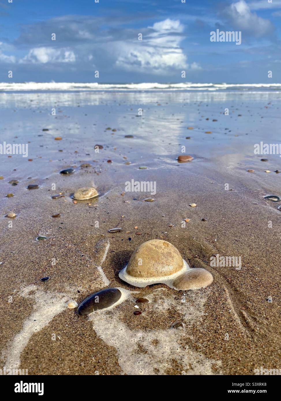 Ciottoli su una spiaggia bagnata dopo che la marea si è ritirata con le onde che si infrangono sullo sfondo e il cielo blu - Immagine stock catturata con smartphone