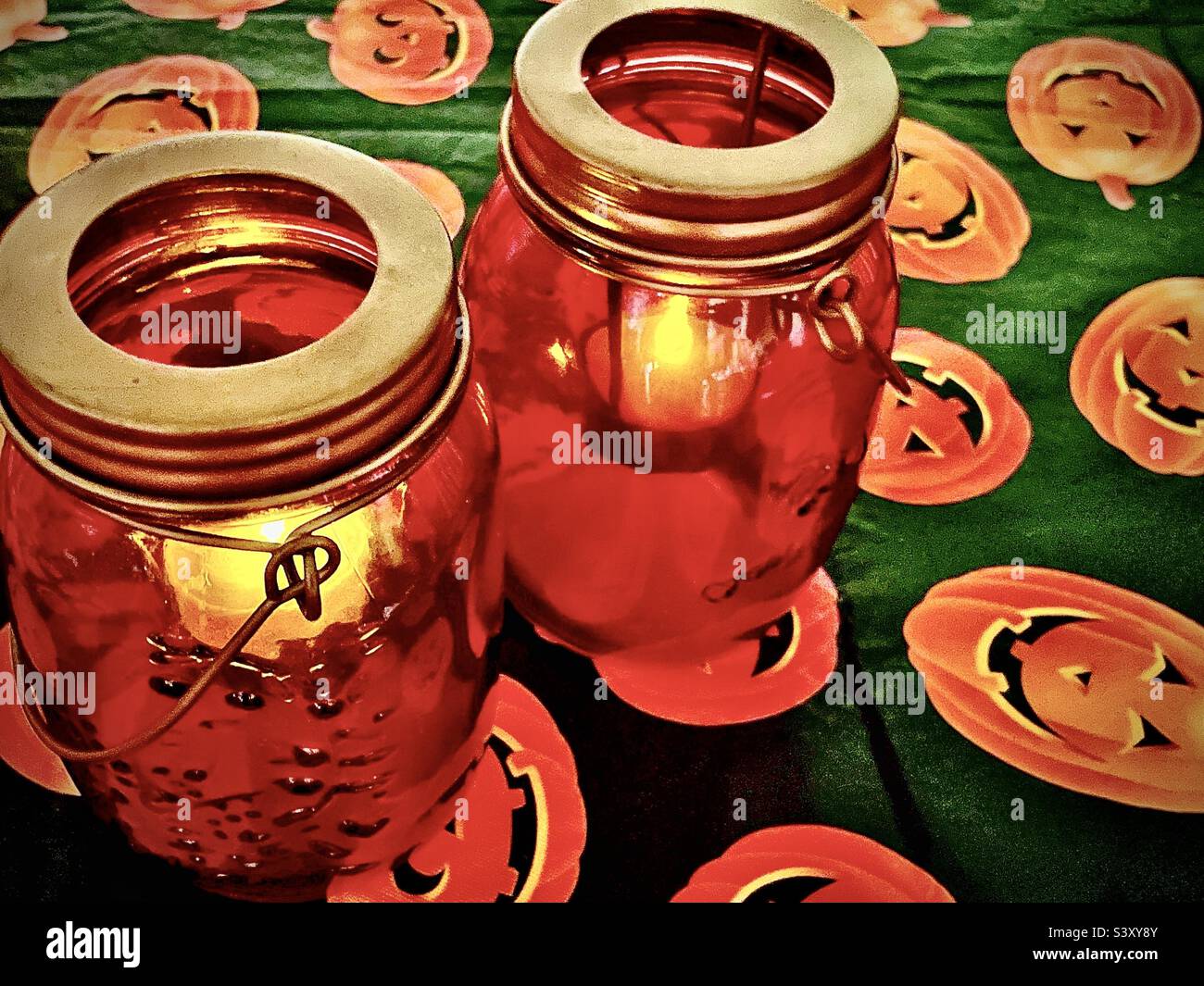 Un paio di candele giara di muratore arancione danno luce tremolante seduto su un tavolo coperto da una tovaglia stagionale di Halloween jack-o-lantern. - Immagine stock catturata con smartphone