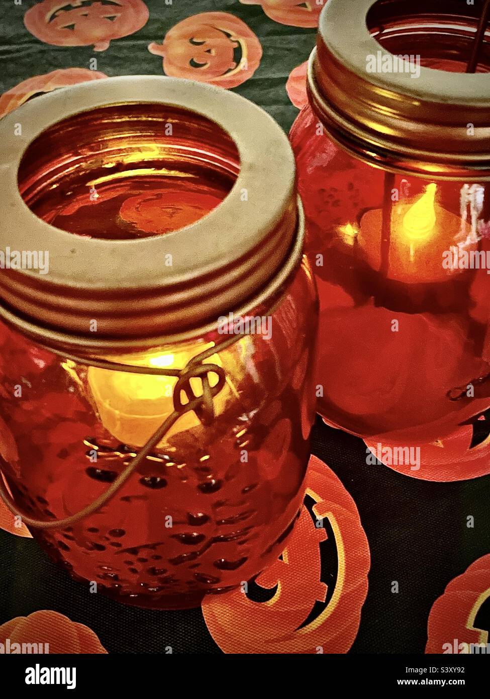 Un paio di candele giara di muratore arancione danno luce tremolante seduto su un tavolo coperto da una tovaglia stagionale di Halloween jack-o-lantern. - Immagine stock catturata con smartphone