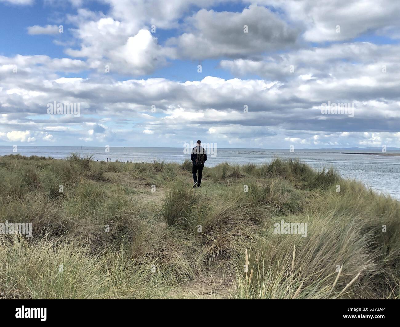 Un uomo si allontana dalla macchina fotografica, tra le erbe soffiate dal vento in un paesaggio costiero, con vista sul mare sullo sfondo e cielo blu con le nuvole sopra. Foto Stock
