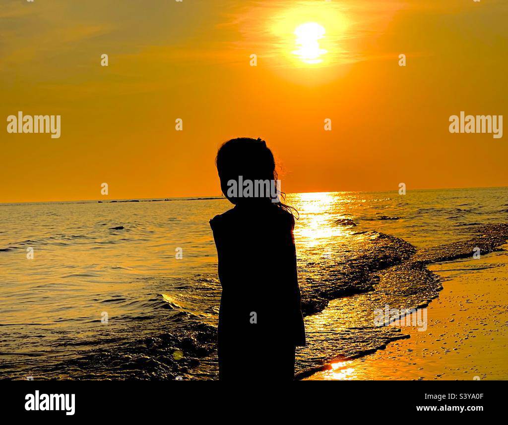 kleines Mädchen am Strand bei Sonnenuntergang das Meer spiegelt in goldenen Farben Foto Stock