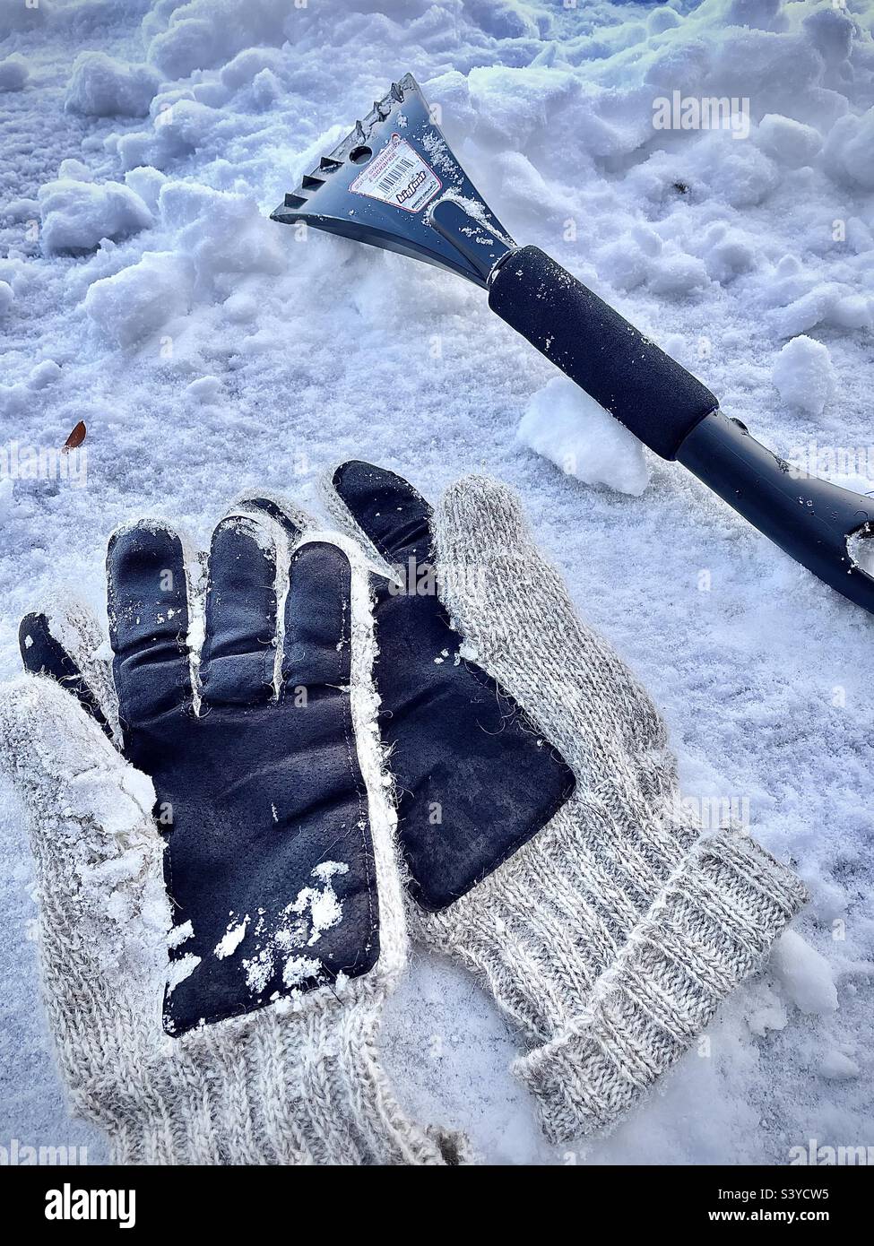 Un paio di guanti di lana e un attrezzo per neve/ghiaccio, un'estremità un raschietto per ghiaccio, l'altra estremità un pennello da neve. Una vita ancora ventosa si è imposta su un cofano di automobile coperto di neve dopo una tempesta di neve dello Utah, Stati Uniti. Foto Stock