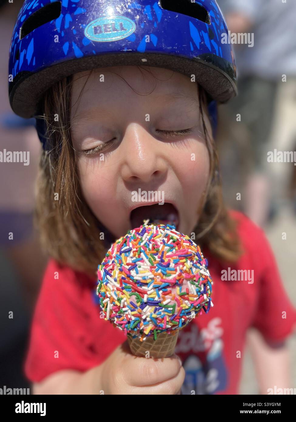 Bambino di cinque anni che indossa un casco da bicicletta e che si gode un cono gelato coperto da spruzzi colorati. - Immagine stock catturata con smartphone