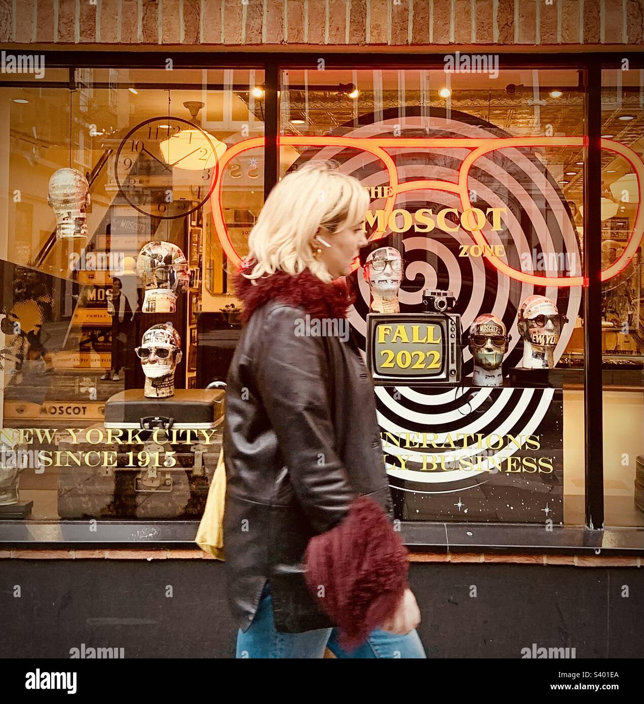 Donna con airpod che cammina davanti a un negozio di occhiali ottici alla moda con teste modello nella finestra, una spirale, e diversi occhiali e sfumature. Autunno/autunno 2022. Moscot a Londra Foto Stock