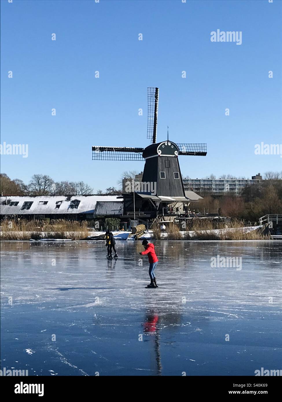 Giovane donna su un cappotto rosso che pattina su un panoramico lago ghiacciato con un mulino a vento sul retro - Immagine stock catturata con smartphone