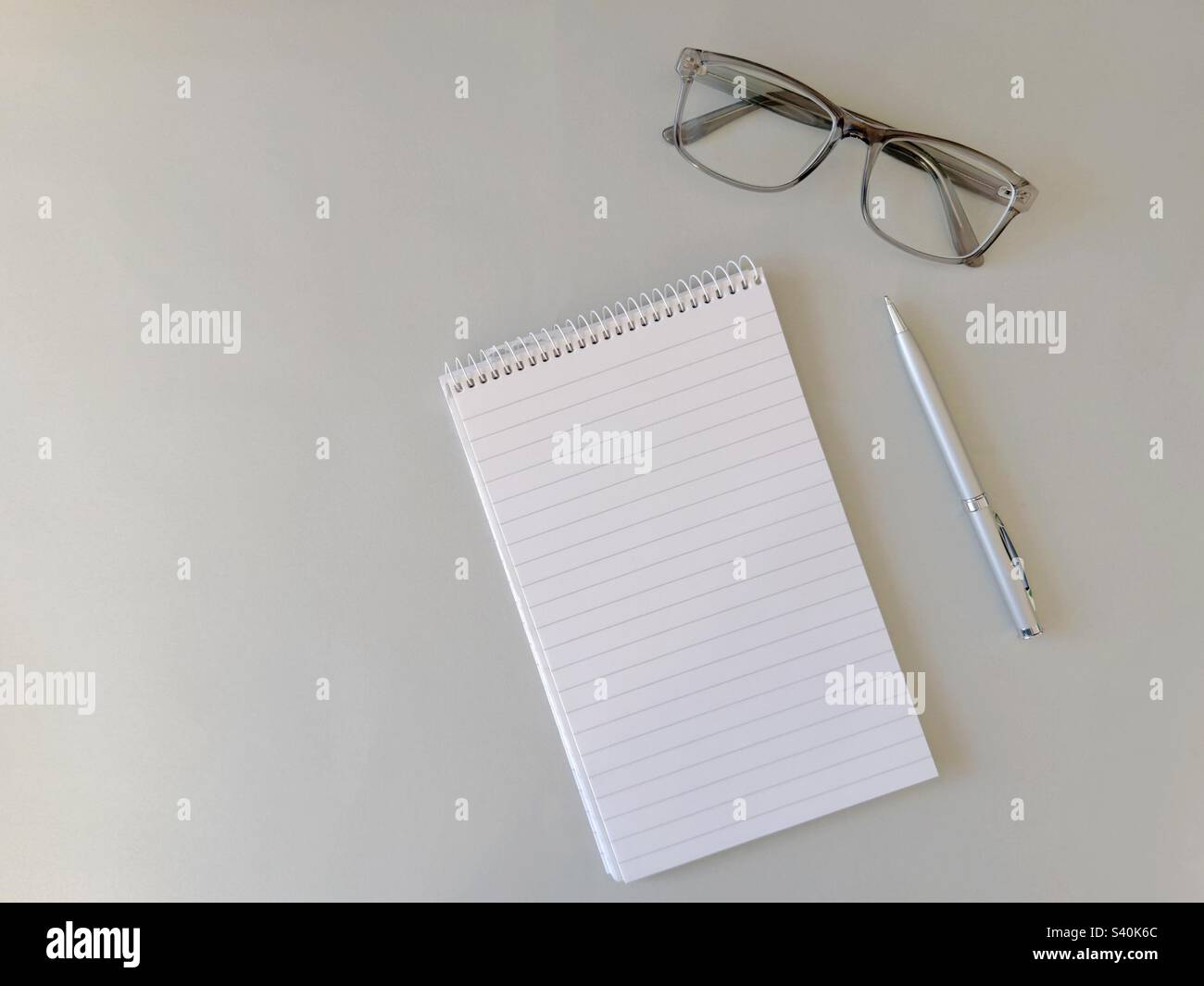 Display piatto di un blocco note e di una penna con un paio di occhiali su uno sfondo grigio chiaro. Spazio di copia. Foto Stock