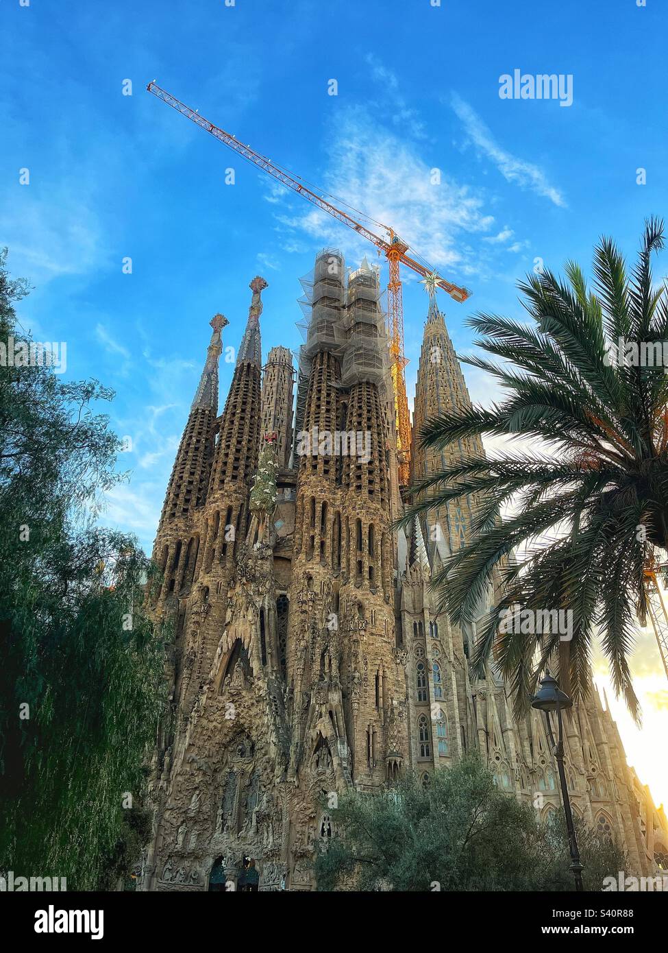 La facciata della Natività di Antoni Gaudí e la Cripta della Basílica de la Sagrada Família, Barcellona, vista attraverso gli alberi del parco locale, Barcellona, Spagna Foto Stock
