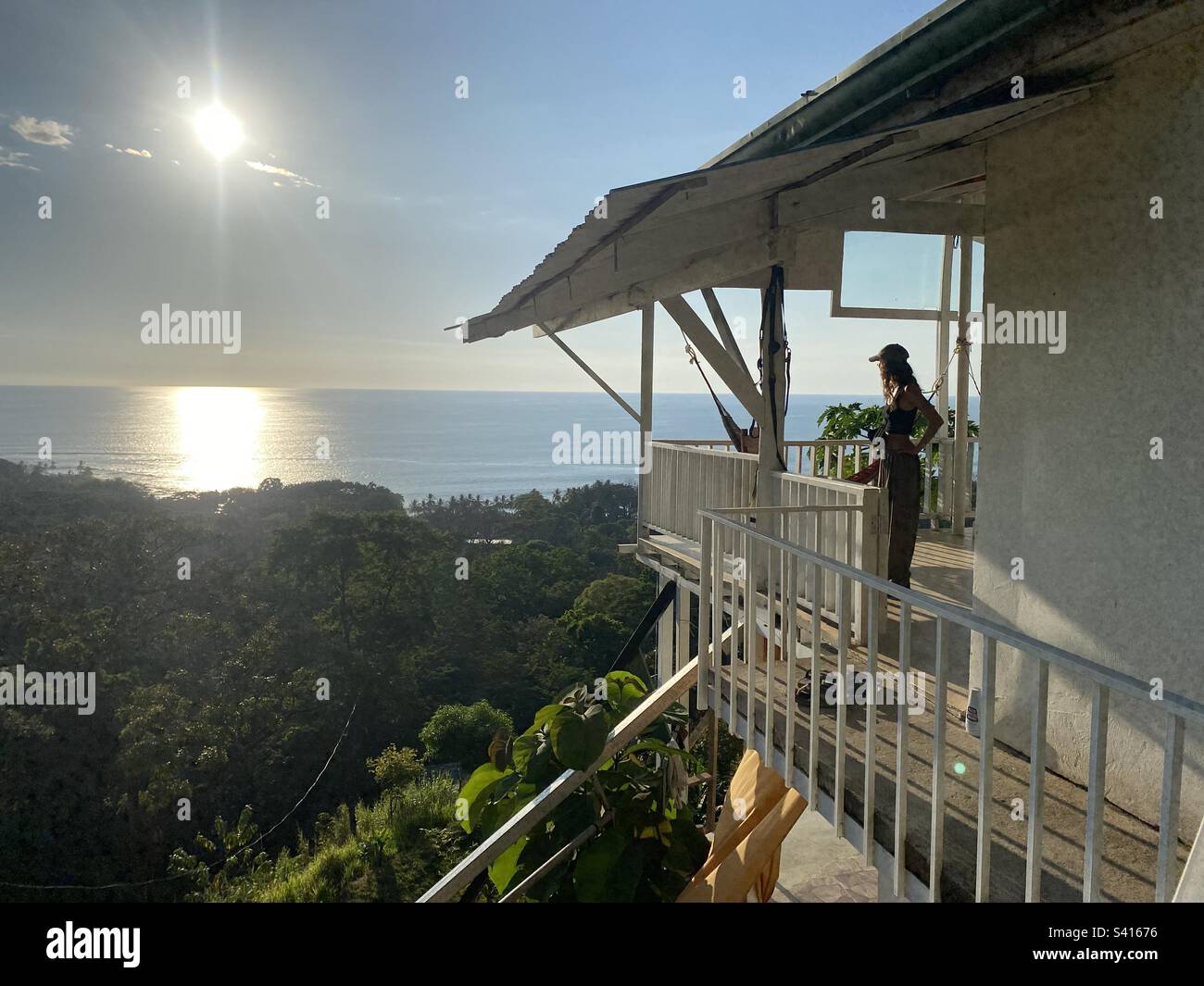 Una donna in piedi sul balcone di una casa in collina con viste spettacolari del mare e del tramonto Foto Stock