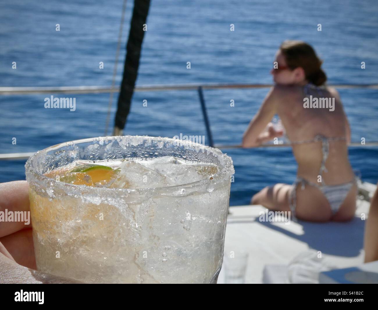 Una donna in un bikini siede sulla parte anteriore di una barca a vela con un drink margarita in primo piano - Immagine stock catturata con smartphone