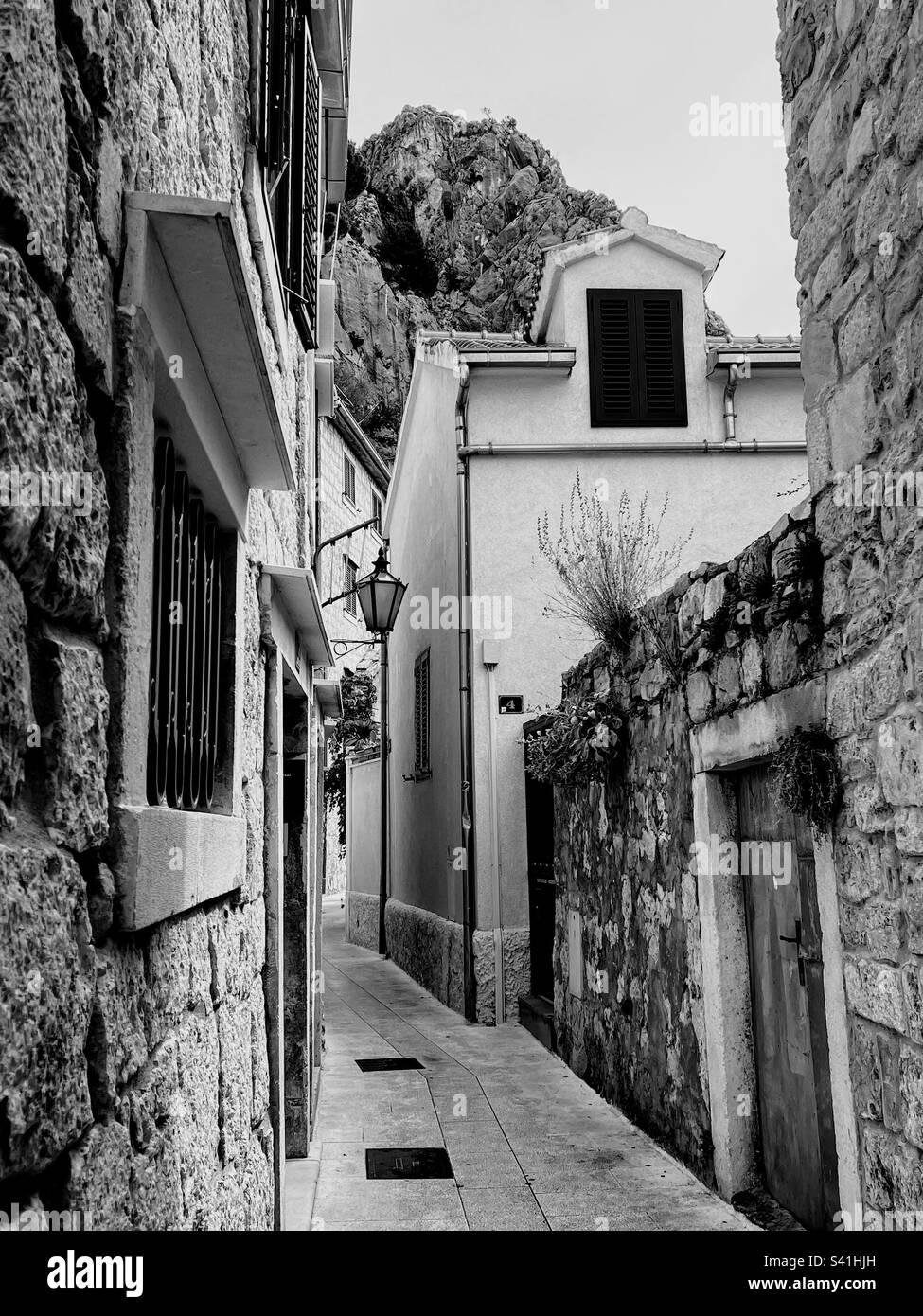 Foto monocromatica in bianco e nero di una vecchia strada sterrata tra edifici in pietra arenaria con alta montagna sullo sfondo - Immagine stock catturata con smartphone