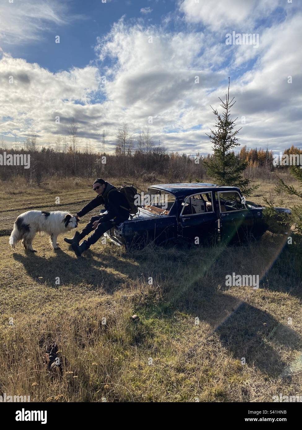 Un uomo con uno zaino è seduto su una vecchia auto abbandonata e accarezzare un cane bianco nella foresta di Yakutia. - Immagine stock catturata con smartphone