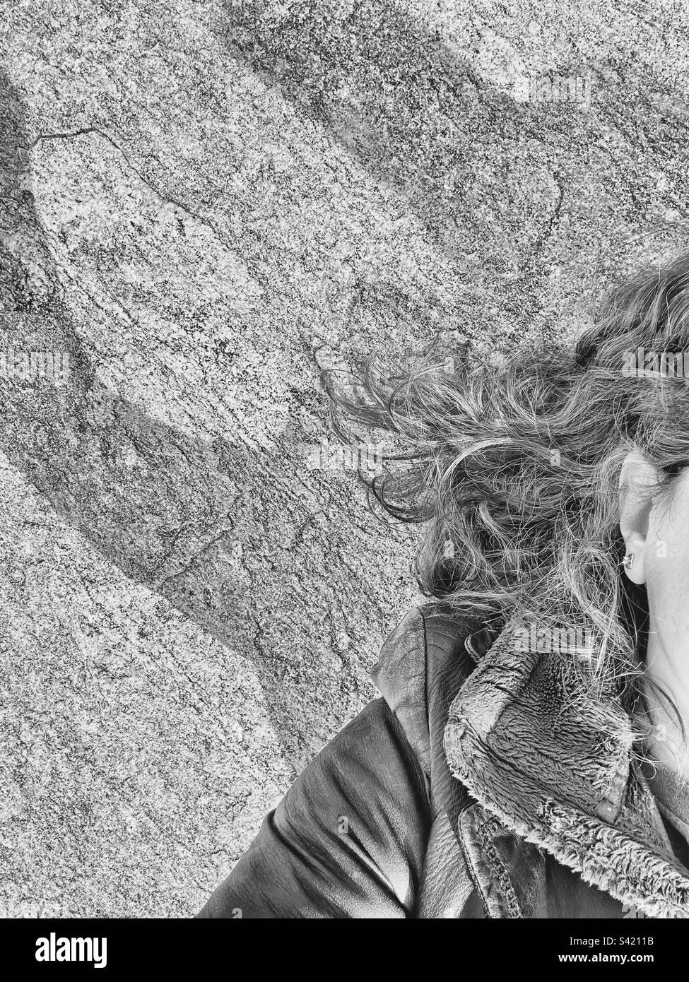 Ritratto di una donna bianca adagiato su una roccia. Mostra solo una piccola parte del viso e i capelli stanno volando fuori. Indossa una giacca in pelle nera e un pullover in pile sotto. Filtro bianco e nero. - Immagine stock catturata con smartphone