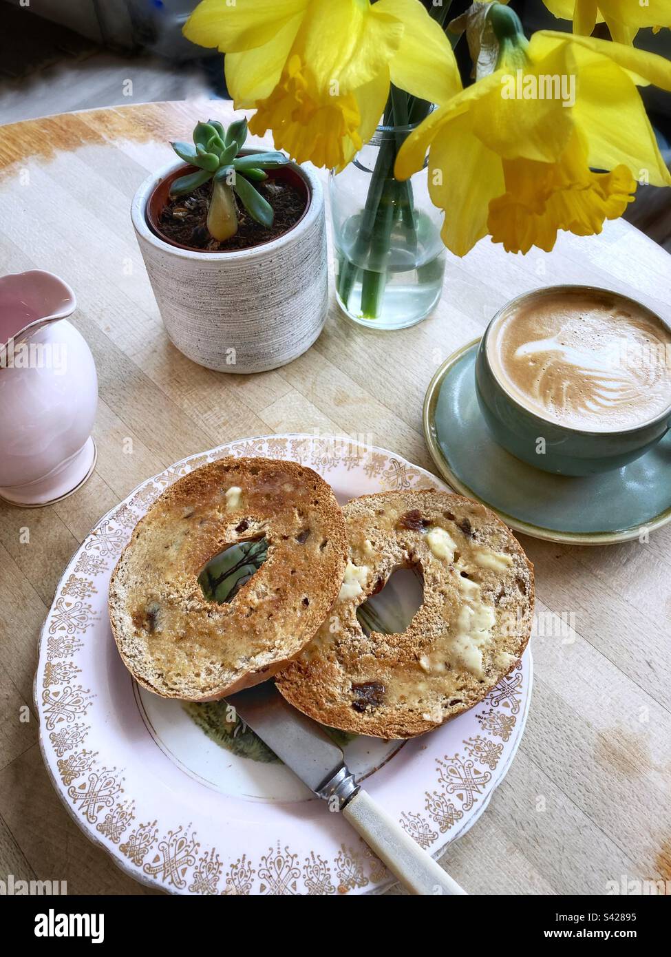 Caffè bianco piatto e un bagel di cannella tostato su un tavolo con le narcisi Foto Stock