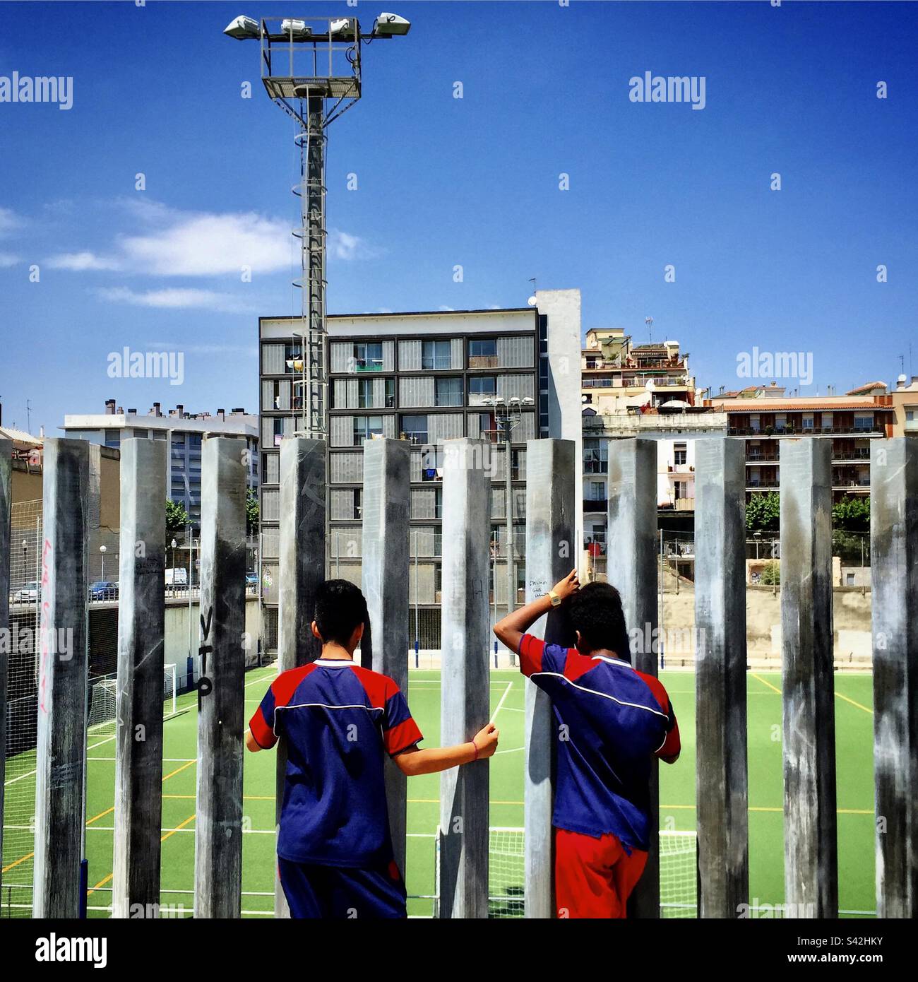 Due ragazzi guardano la partita di calcio allenarsi attraverso le lacune in una recinzione di ferro nel centro di Barcellona Foto Stock