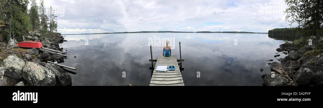 Panorama di una giovane ragazza sedette su un molo che fissava un grande lago in una casa estiva nella regione di Kajaani in Finlandia durante un'estate artica Foto Stock