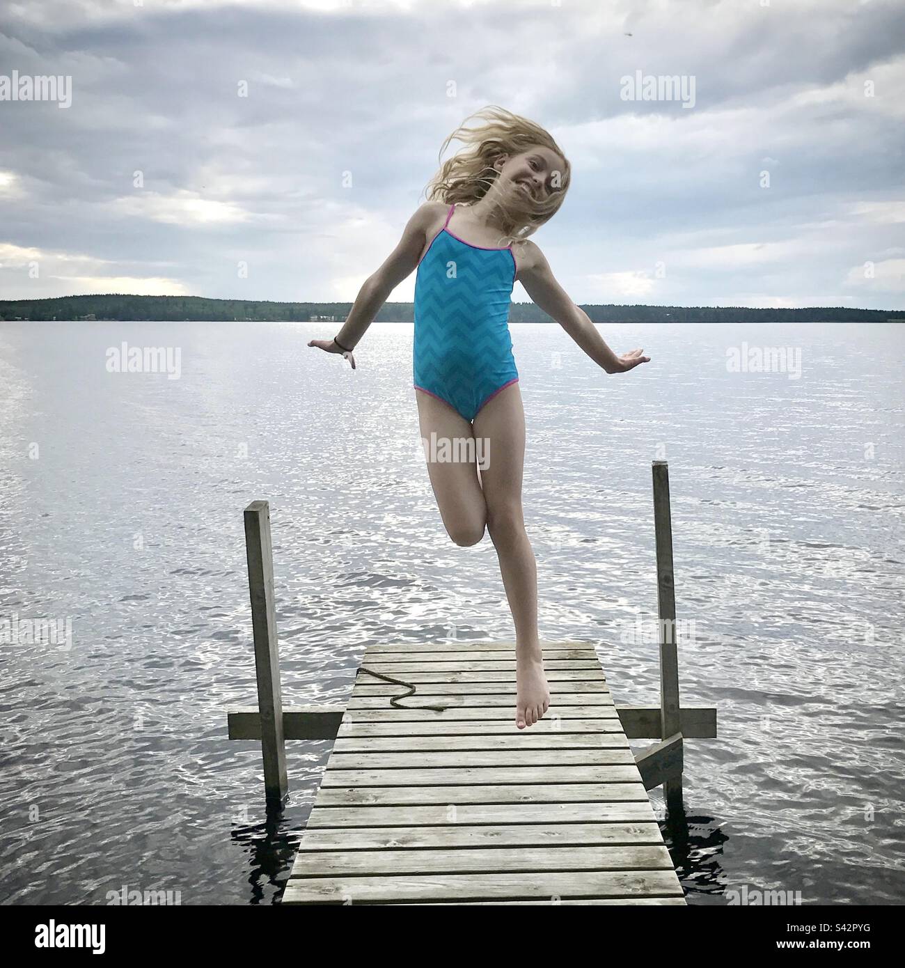 Una giovane ragazza che salta per la gioia su un molo in un grande lago in una casa estiva nella regione di Kajaani in Finlandia durante un'estate artica Foto Stock