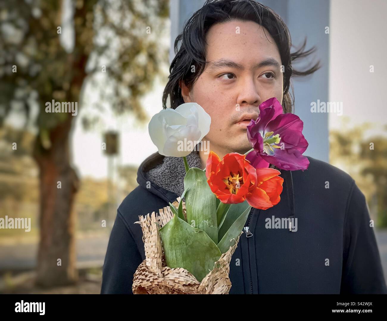 Ritratto di giovane asiatico che tiene bouquet di tulipani contro gli alberi durante il tramonto. - Immagine stock catturata con smartphone