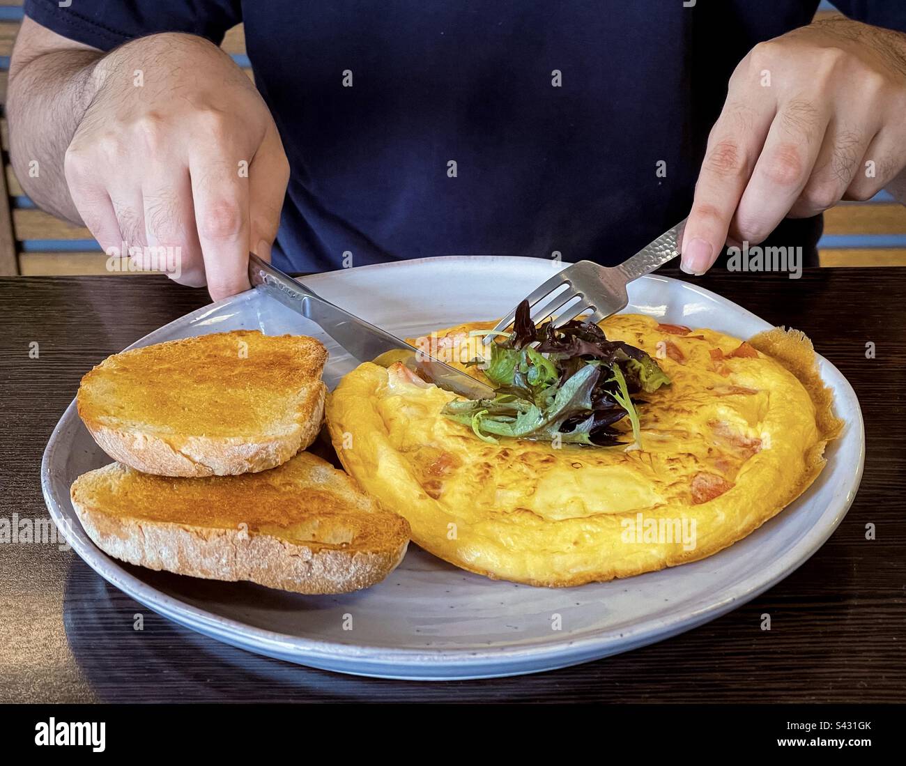 A metà dell'uomo si usano le posate per tagliare l'omelette al formaggio vegetariano sul piatto con il pane a pasta madre. Foto Stock