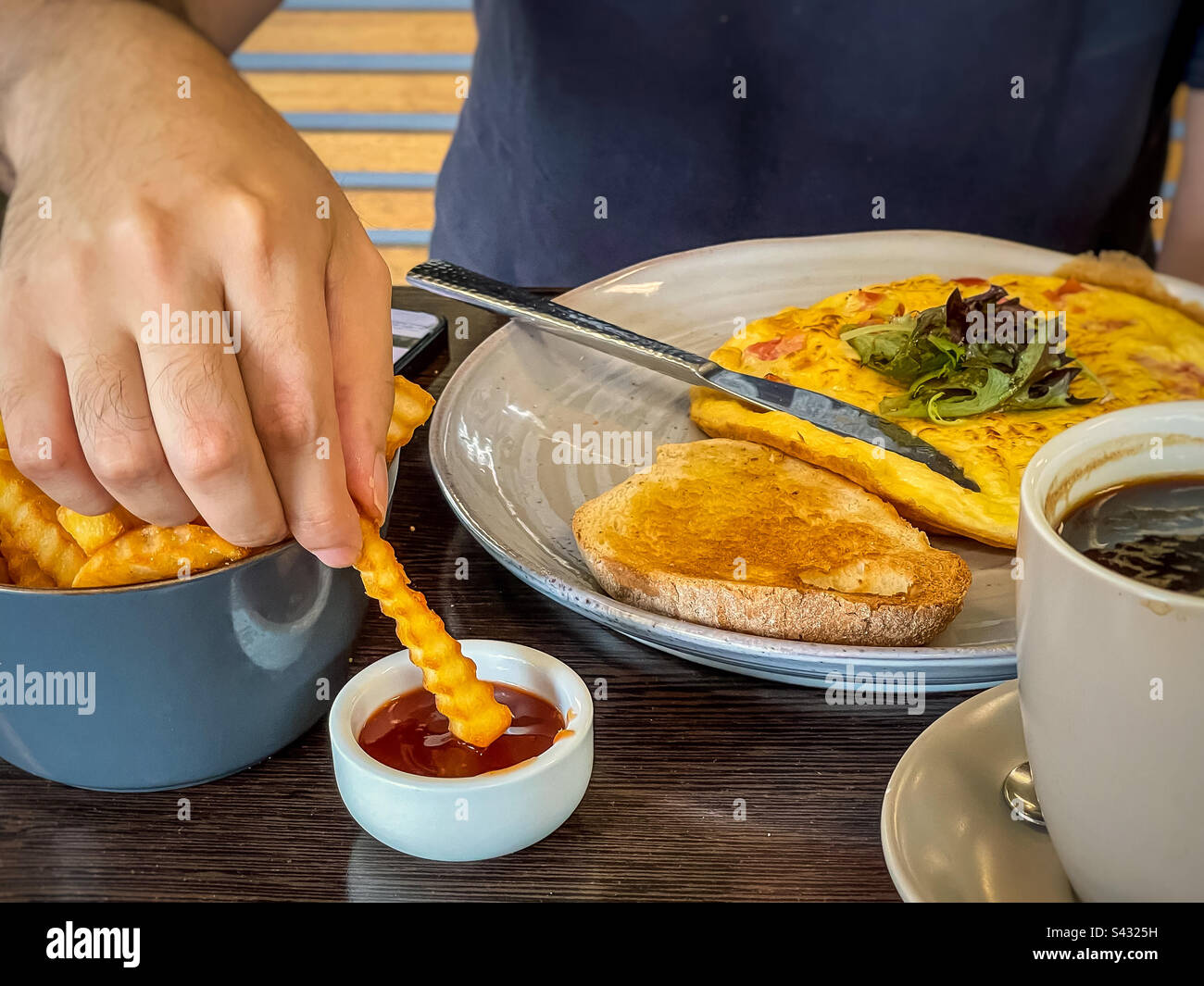 Parte centrale dell'uomo che immerge le patatine fritte in ketchup contro l'omelette e il pane di pasta madre sul piatto. Foto Stock
