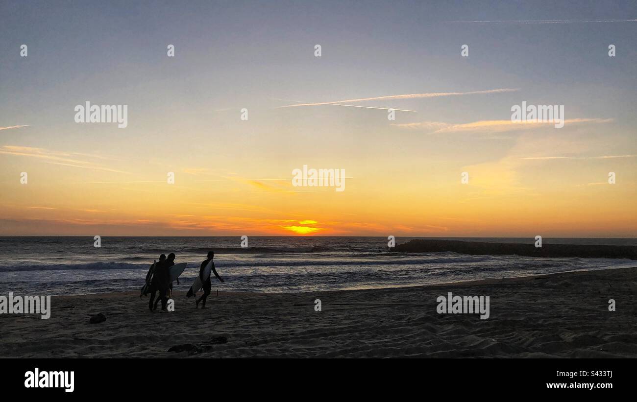 Silouettes di tre surfisti contro il cielo tramonto a piedi lungo la spiaggia di Espinho, Portogallo, 2023 Foto Stock