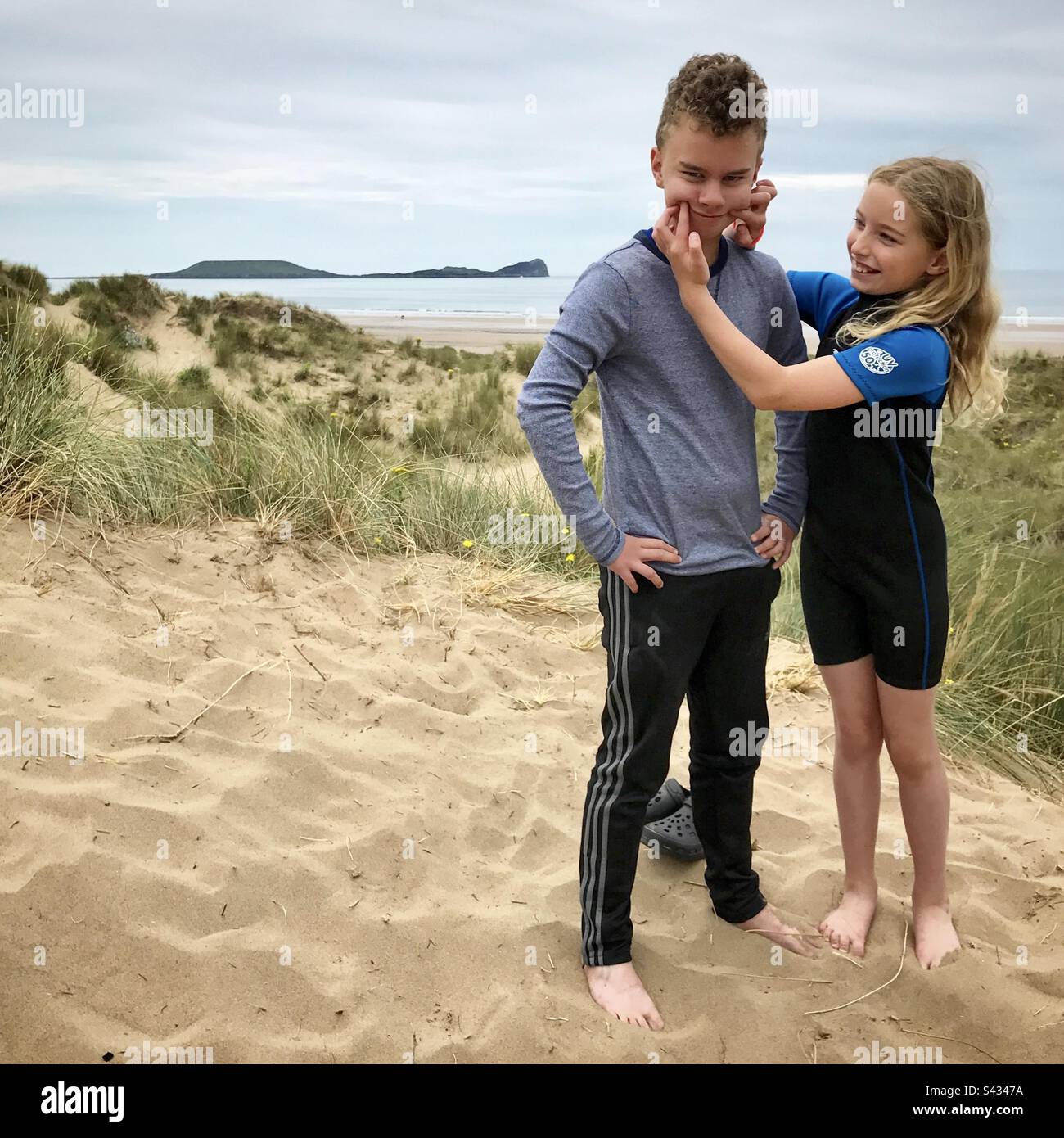 Fratello e sorella che si divertono mentre la giovane ragazza cerca di far sorridere il ragazzo in una duna di sabbia sulla spiaggia di Rhossili nel Galles occidentale Foto Stock