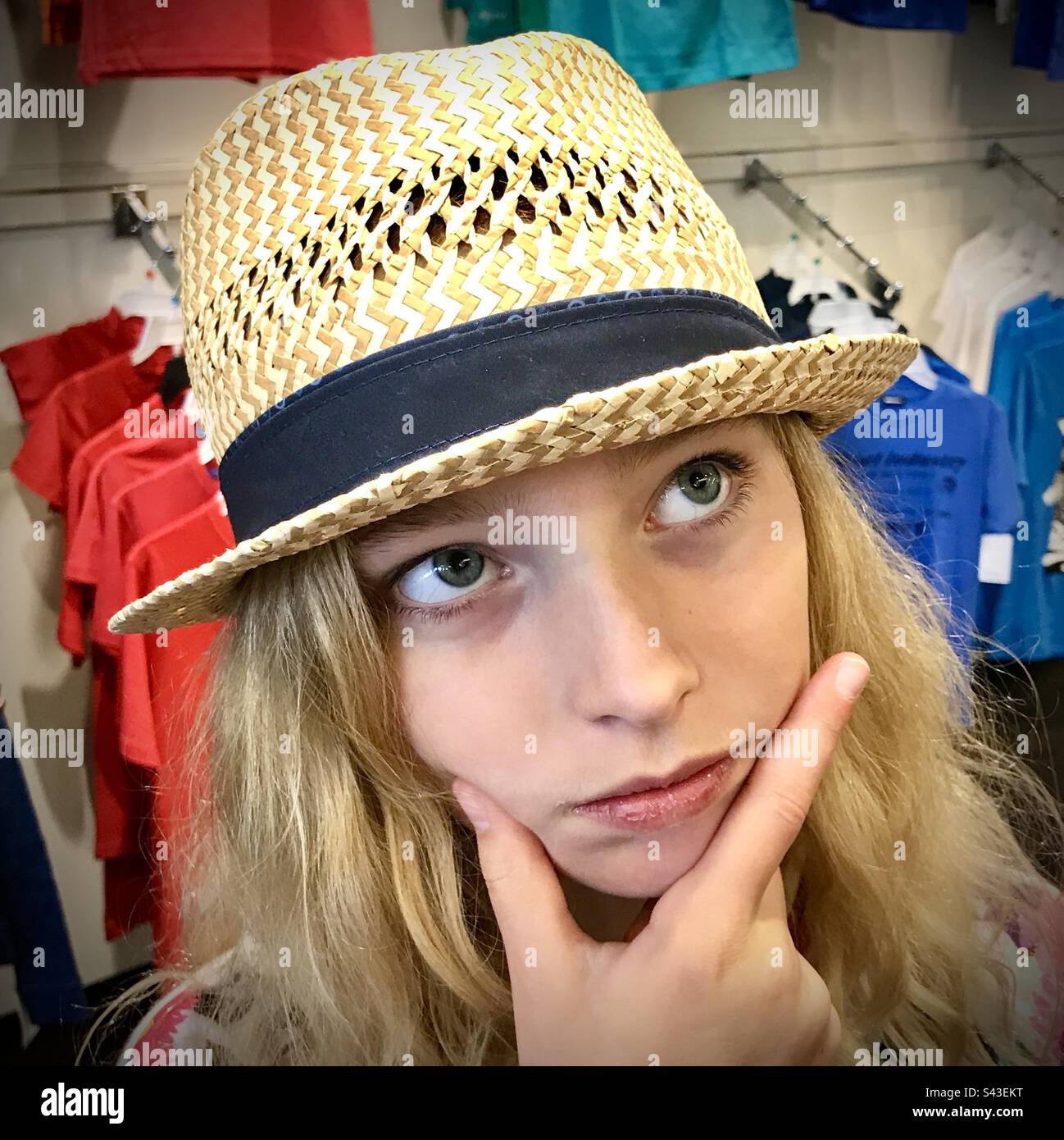 Una giovane ragazza di 10 anni che fa shopping recita guardando confuso mentre si acquista in un cappello di paglia oversize Foto Stock