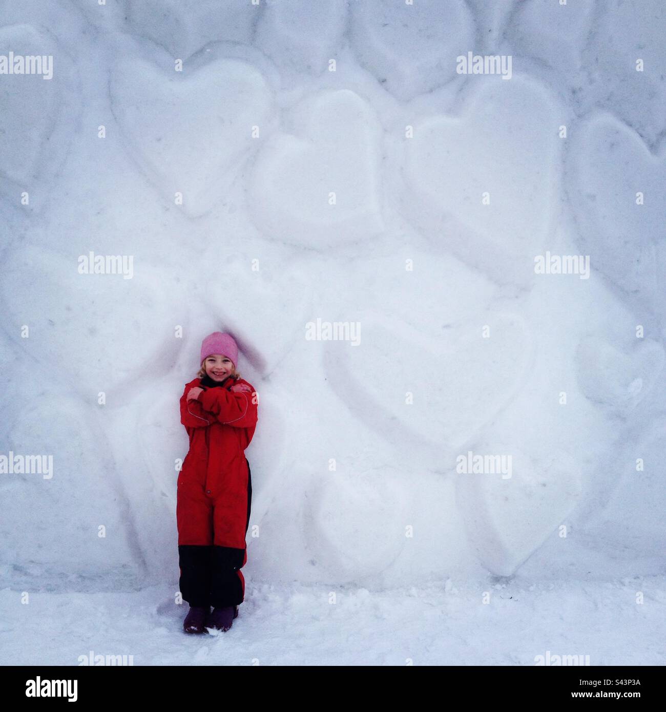 Una giovane ragazza di 6 anni di fronte ad una parete del cuore innevato in un parco giochi pubblico nel profondo inverno in Scandinavia Foto Stock