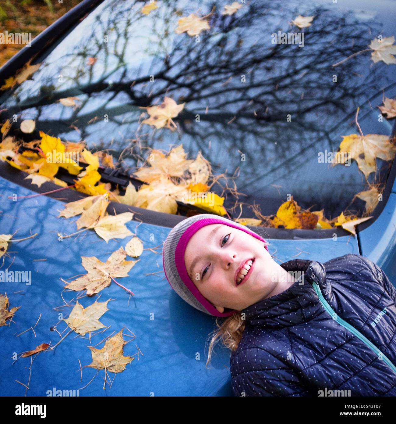 Giovane ragazza di 8 anni che posa sul cofano di un'auto coperta in autunno lascia sorridere mentre le stagioni cambiano l'estate si trasforma in autunno Foto Stock