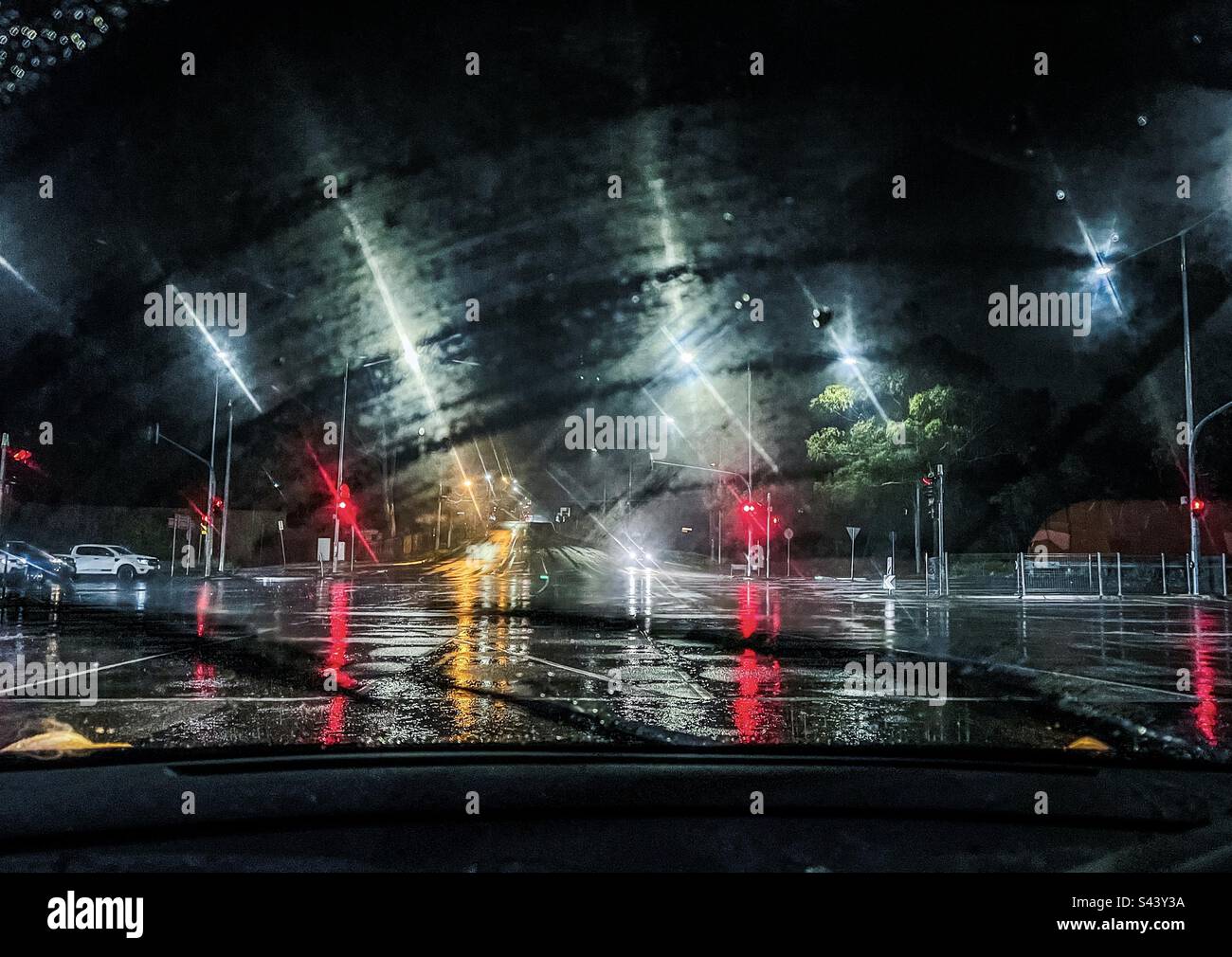 Vista dell'incrocio stradale, dei semafori, delle automobili e dei semafori visti attraverso il parabrezza dell'auto in una notte piovosa. - Immagine stock catturata con smartphone