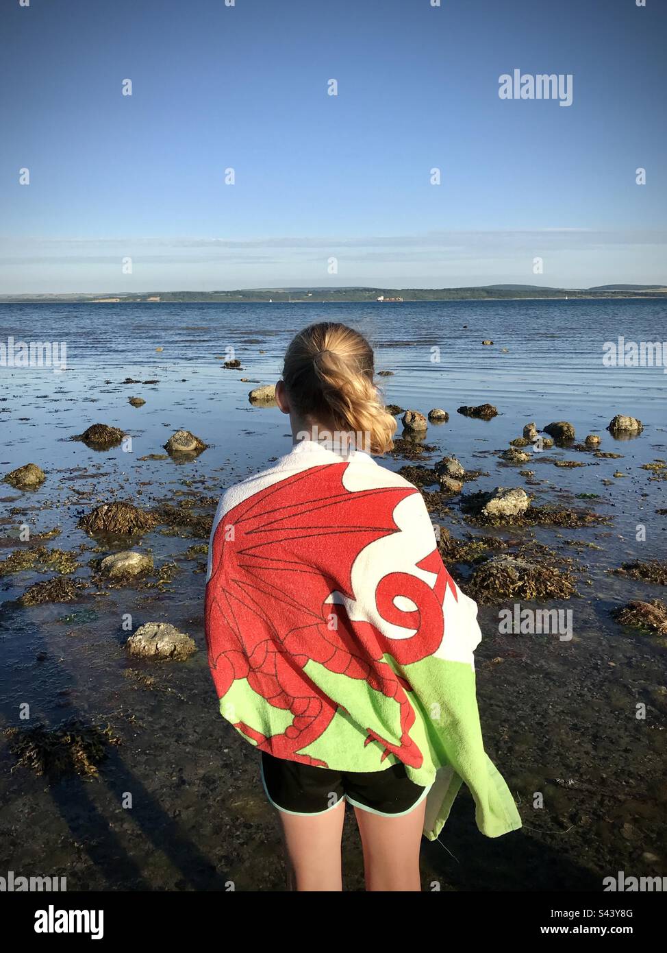 Una ragazza di 10 anni con ritorno alla macchina fotografica avvolta in un asciugamano Wales Flag che si affaccia sul mare all'Isola di Wight da una spiaggia sulla costa meridionale dell'Inghilterra Foto Stock