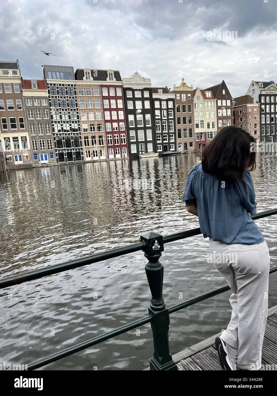 Donne con camicia blu e pantaloni bianchi in piedi sul ponte di Amsterdam, Paesi Bassi, di fronte al fiume, e ci sono acqua casa sullo sfondo Foto Stock