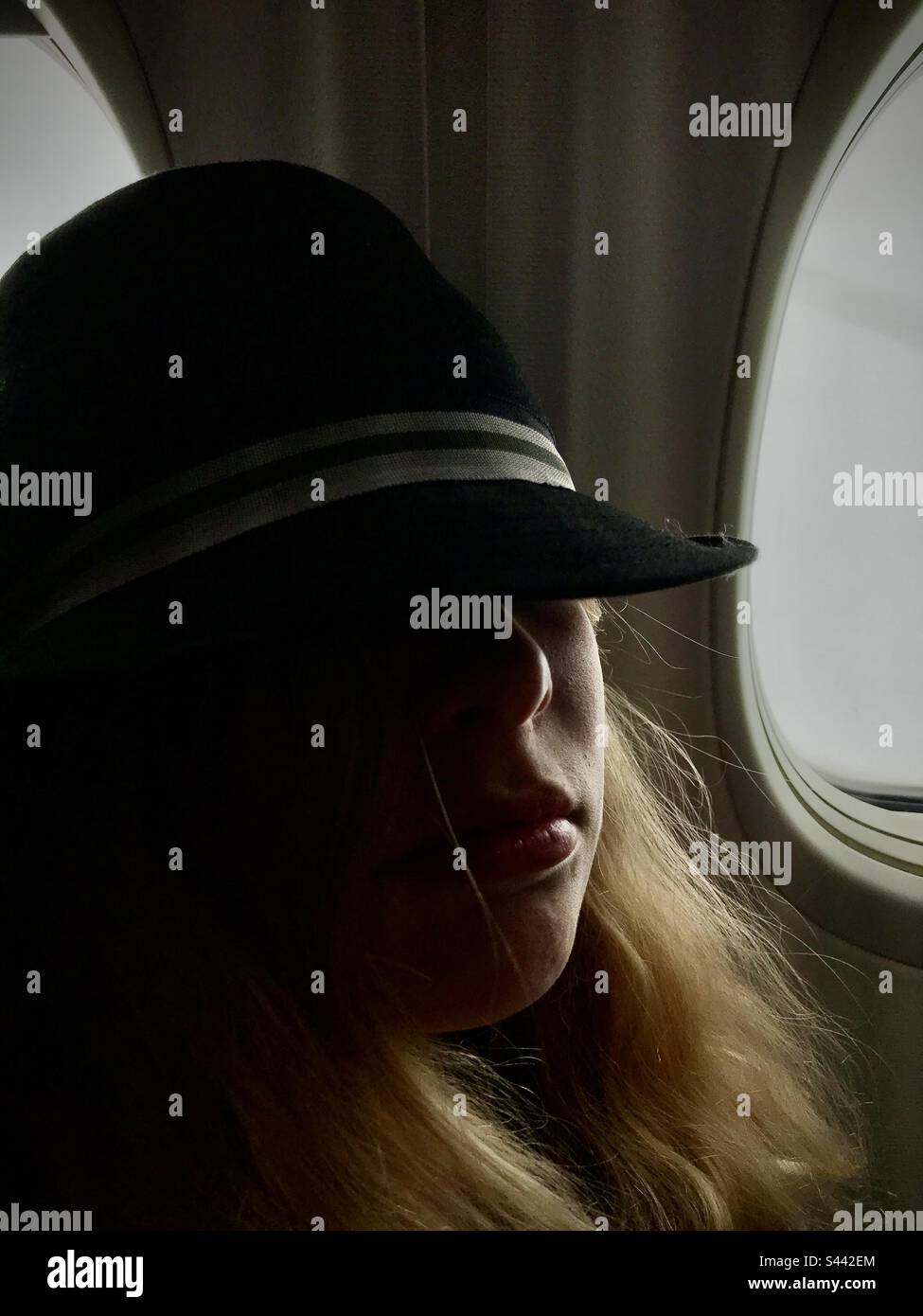 Una giovane ragazza adolescente di 11 anni esausta a dormire prendendo una siesta su un aereo aereo volo cercando fresco sotto un cappello seduto in una finestra seduta in silhouette Foto Stock