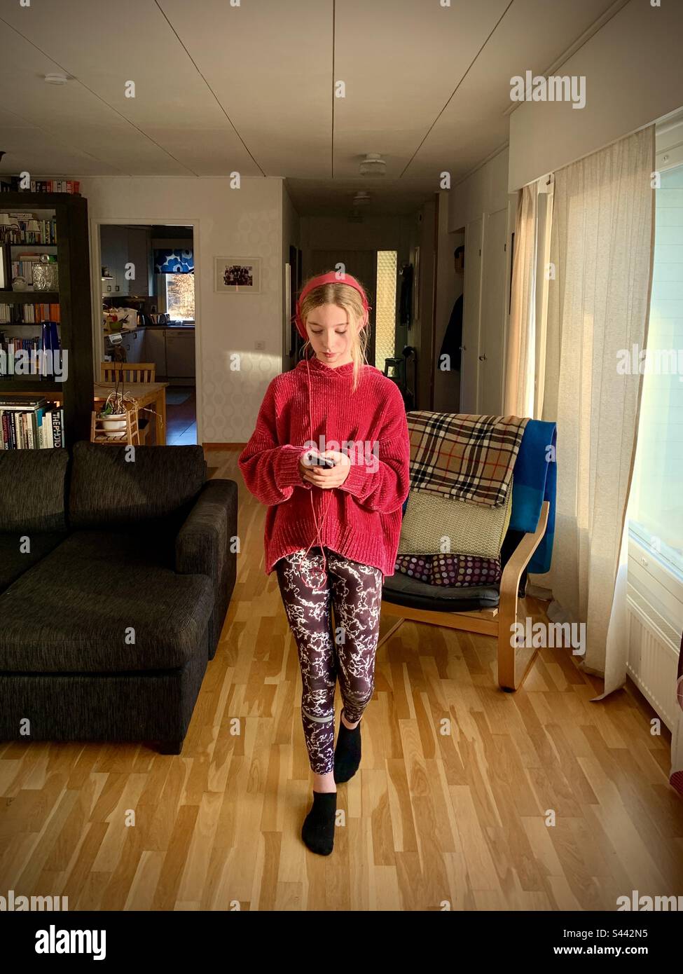 Giovane ragazza adolescente di 11 anni in cuffie rosa scegliendo brani sul suo cellulare playlist profondo nel pensiero perso in musica a piedi attraverso il suo salotto a casa Foto Stock