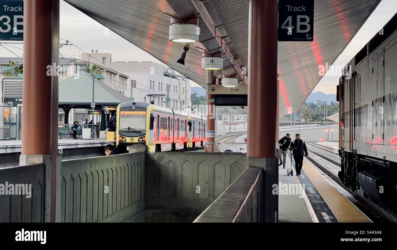 LOS ANGELES, CA, DEC 2022: Due passeggeri che si avvicinano al lato di un treno Metrolink, utilizzando scooter sul binario alla Union Station nel centro. Treno LA Metro Gold Line visibile sullo sfondo Foto Stock